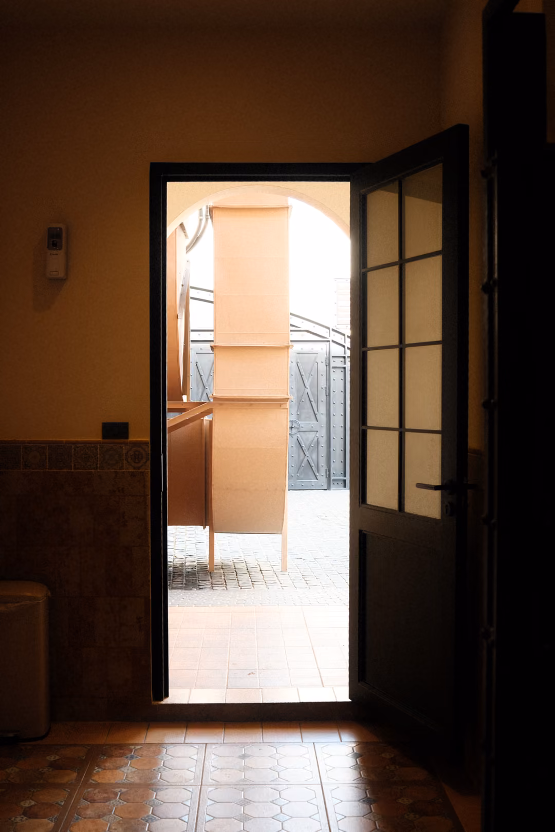 Open dark-framed glass door leading from a dim tiled room to a bright arched passage outside