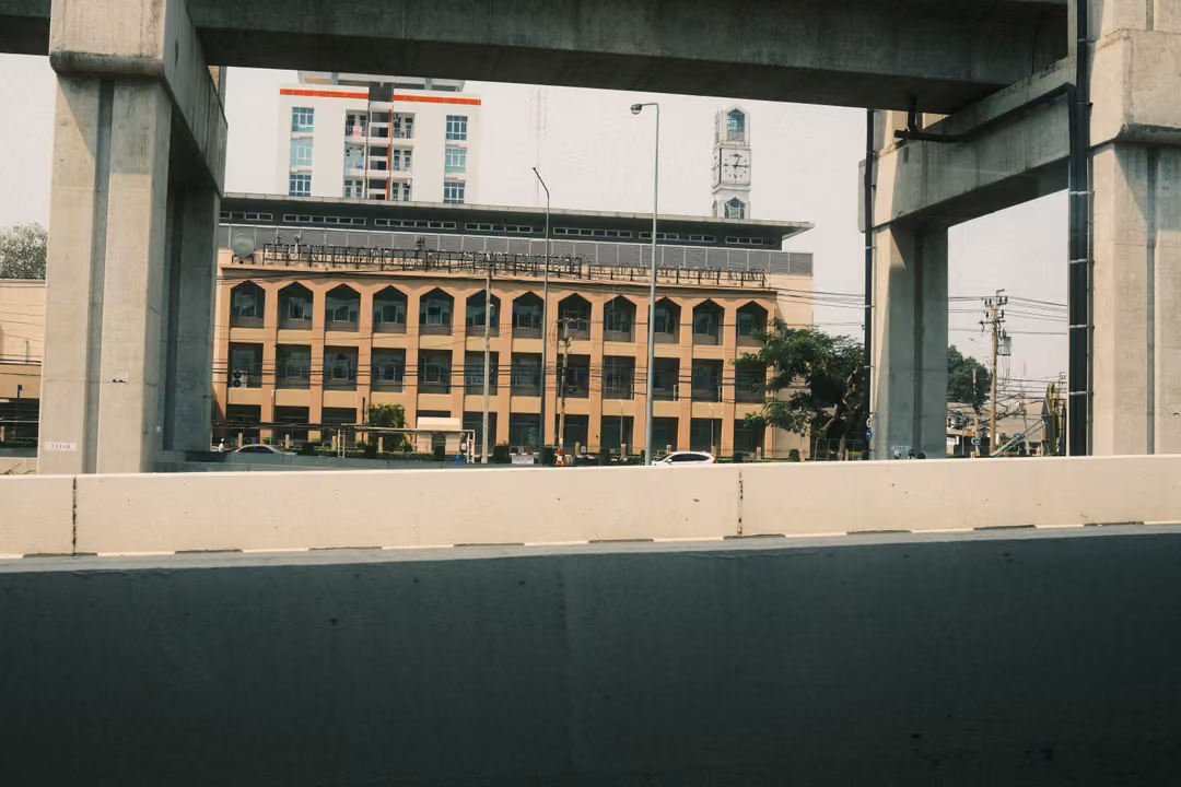 Cream building with arched windows framed between concrete pillars and an elevated track, a clock tower behind