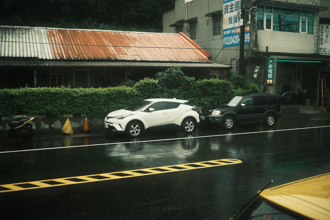 Rainy street seen from a vehicle, a white and a black car parked by a hedge under a rusted corrugated roof
