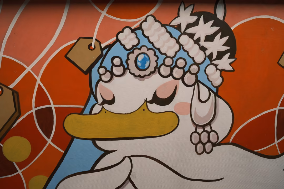 Mural of a white duck-like character with eyes closed, wearing a decorated blue headdress, on an orange background