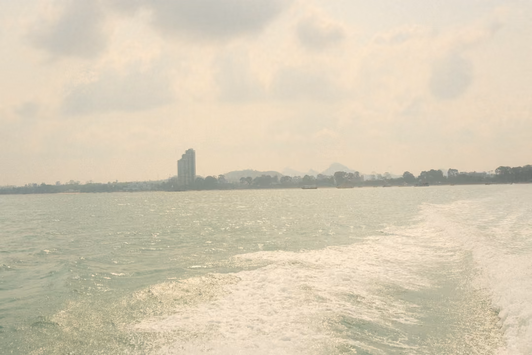 A broad white wake spreads across green sea toward a hazy shore with a single tall tower.