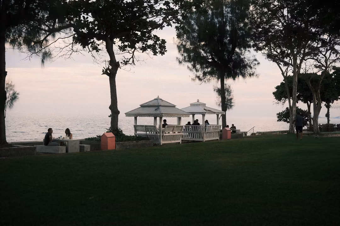 Silhouetted trees above white gazebos and benches by the sea, people sitting along the shore in evening light
