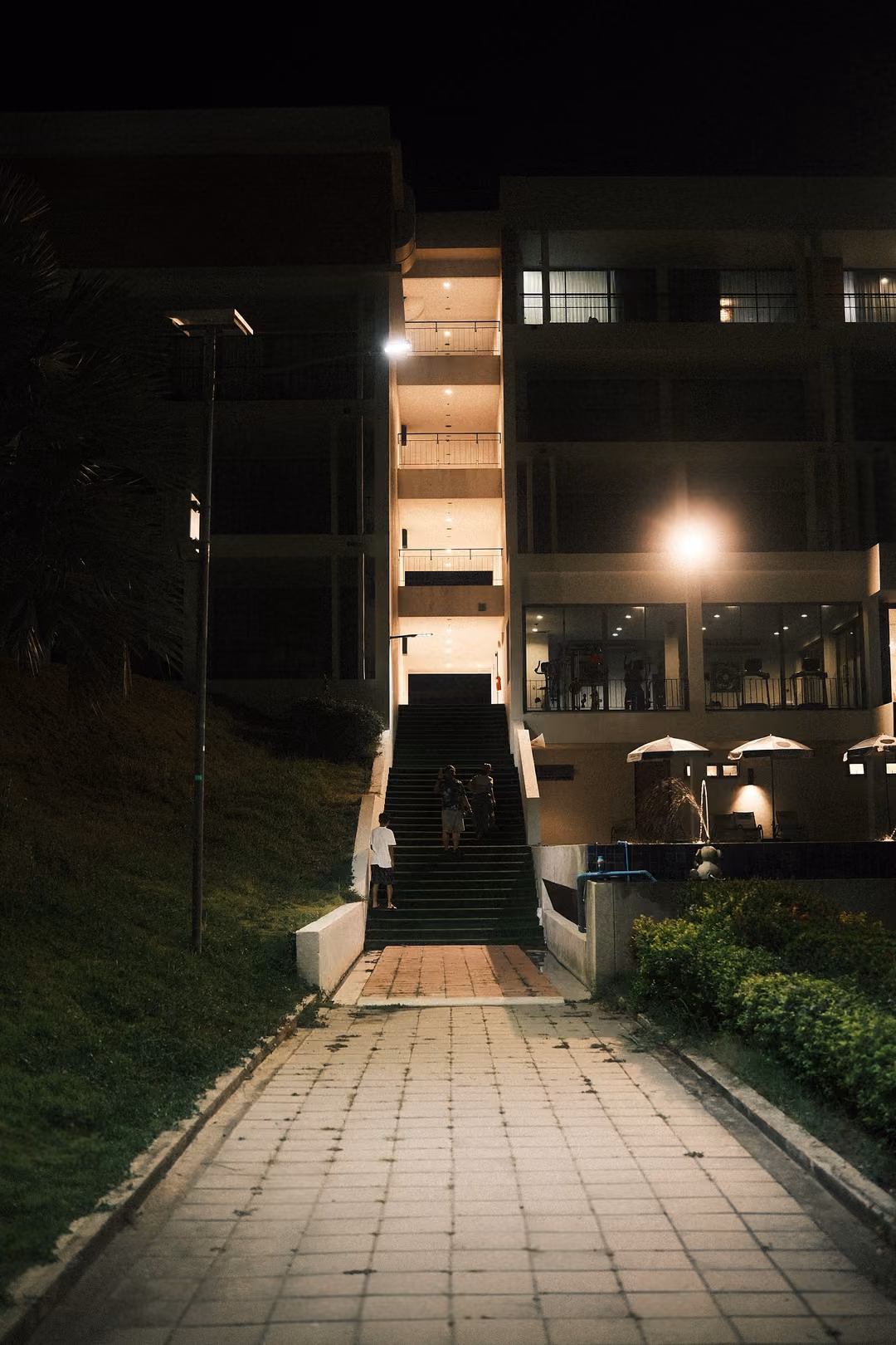 A lit paved walkway and stairs leading up through a grassy slope to a tall hotel building at night