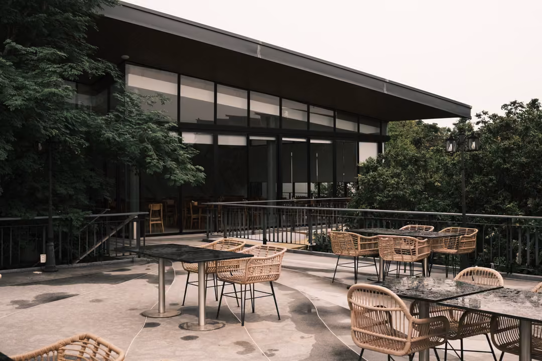 An empty outdoor terrace with wicker chairs and dark tables beside a modern glass-fronted building.