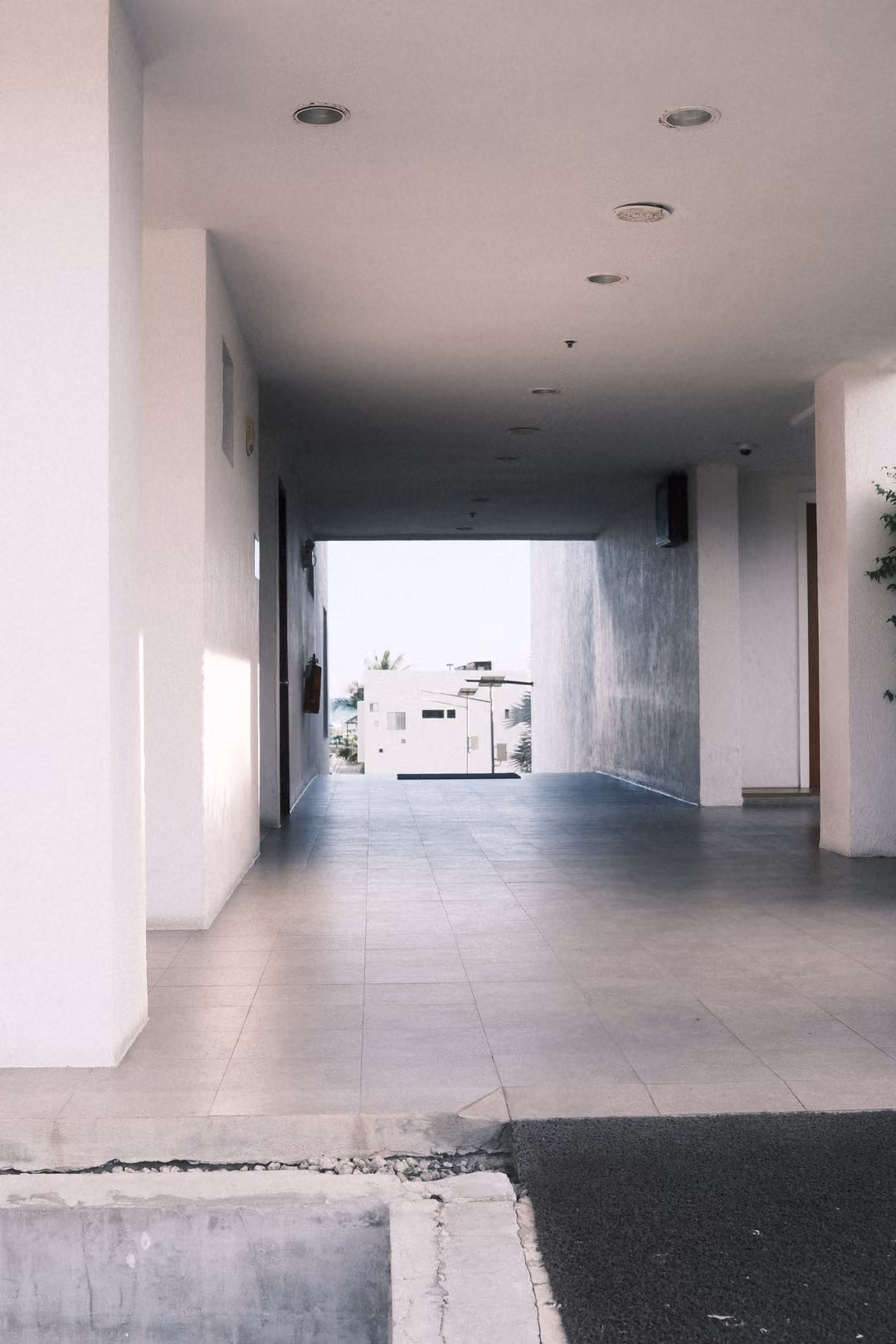 Covered white walkway with recessed ceiling lights leading toward a bright exit and a white building outside