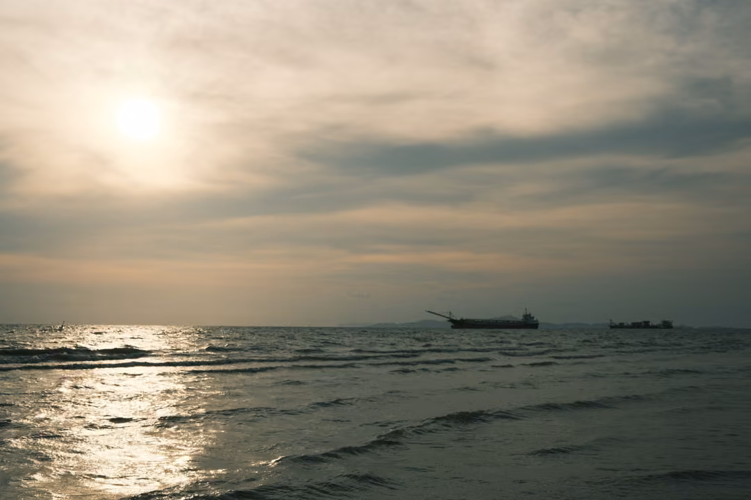 A cloudy sky over the sea with a soft sun, a ship with a long crane on the horizon