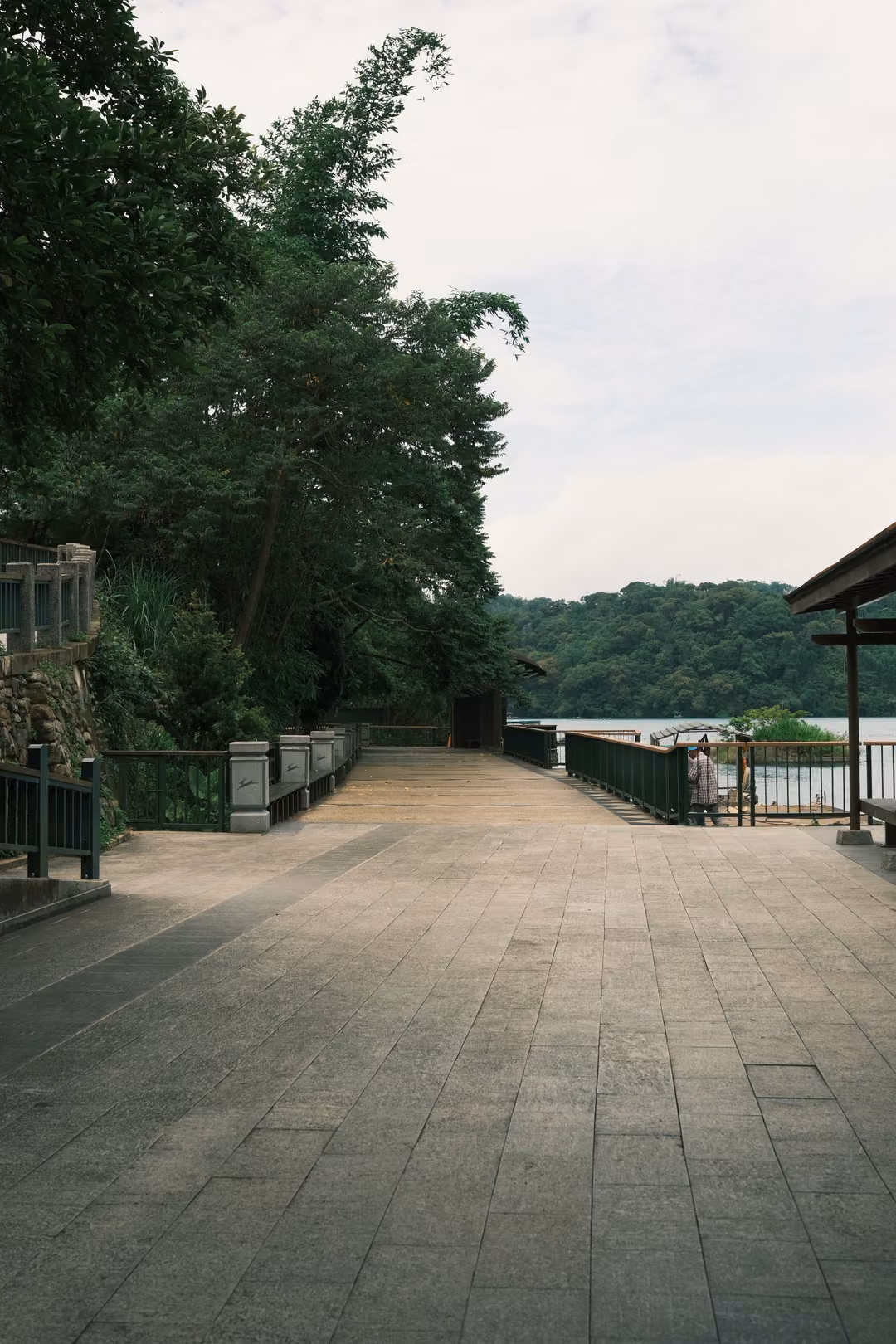 Wide paved lakeside path under trees and a pavilion roof, railings and water visible in the distance