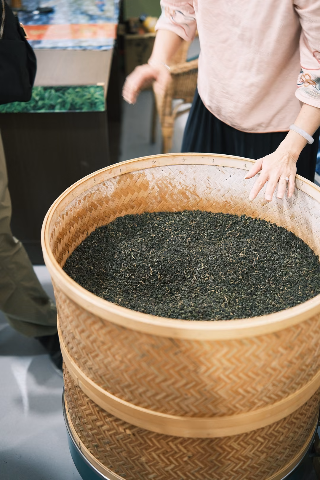 Hand resting on the rim of a large woven bamboo basket filled with dark rolled tea leaves