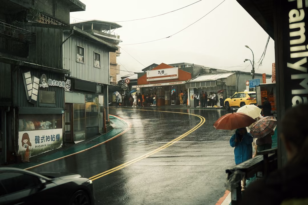 Rainy curving street with umbrellas, a yellow taxi and shopfronts, a FamilyMart sign at the right edge