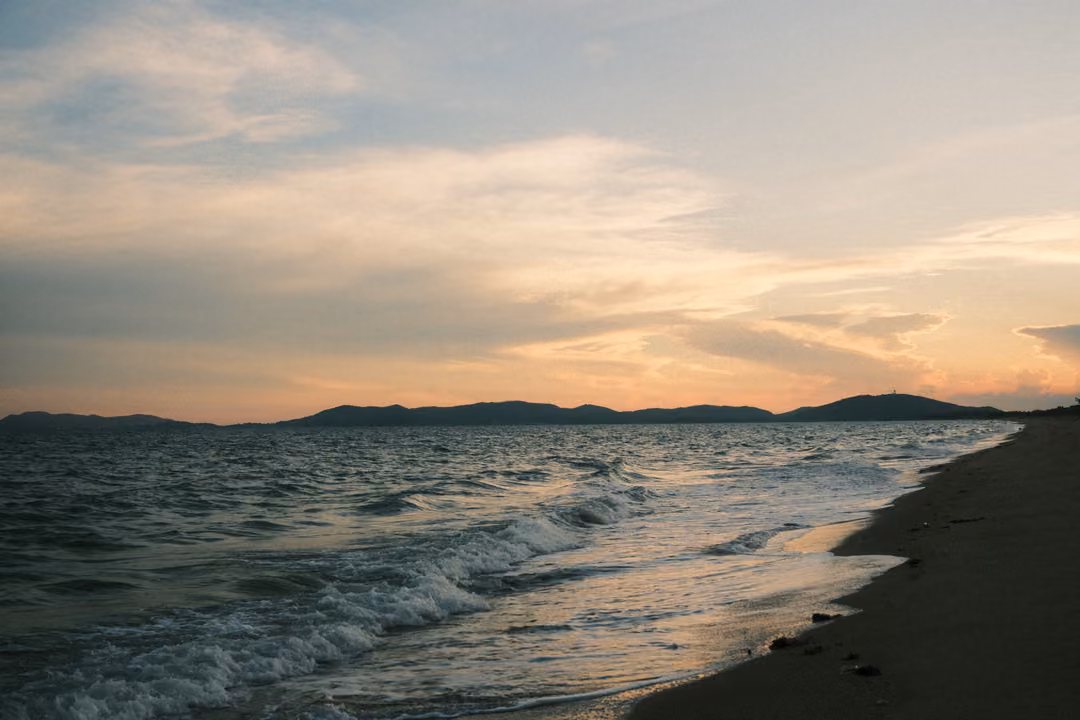 Waves wash up a curving sandy shore at sunset, the sky pink and blue, dark hills on the horizon