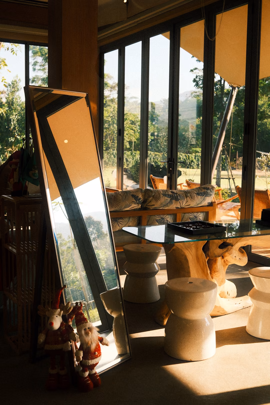 Tall leaning mirror reflecting a sunlit room, small Santa figurines at its base, white stools and a glass table