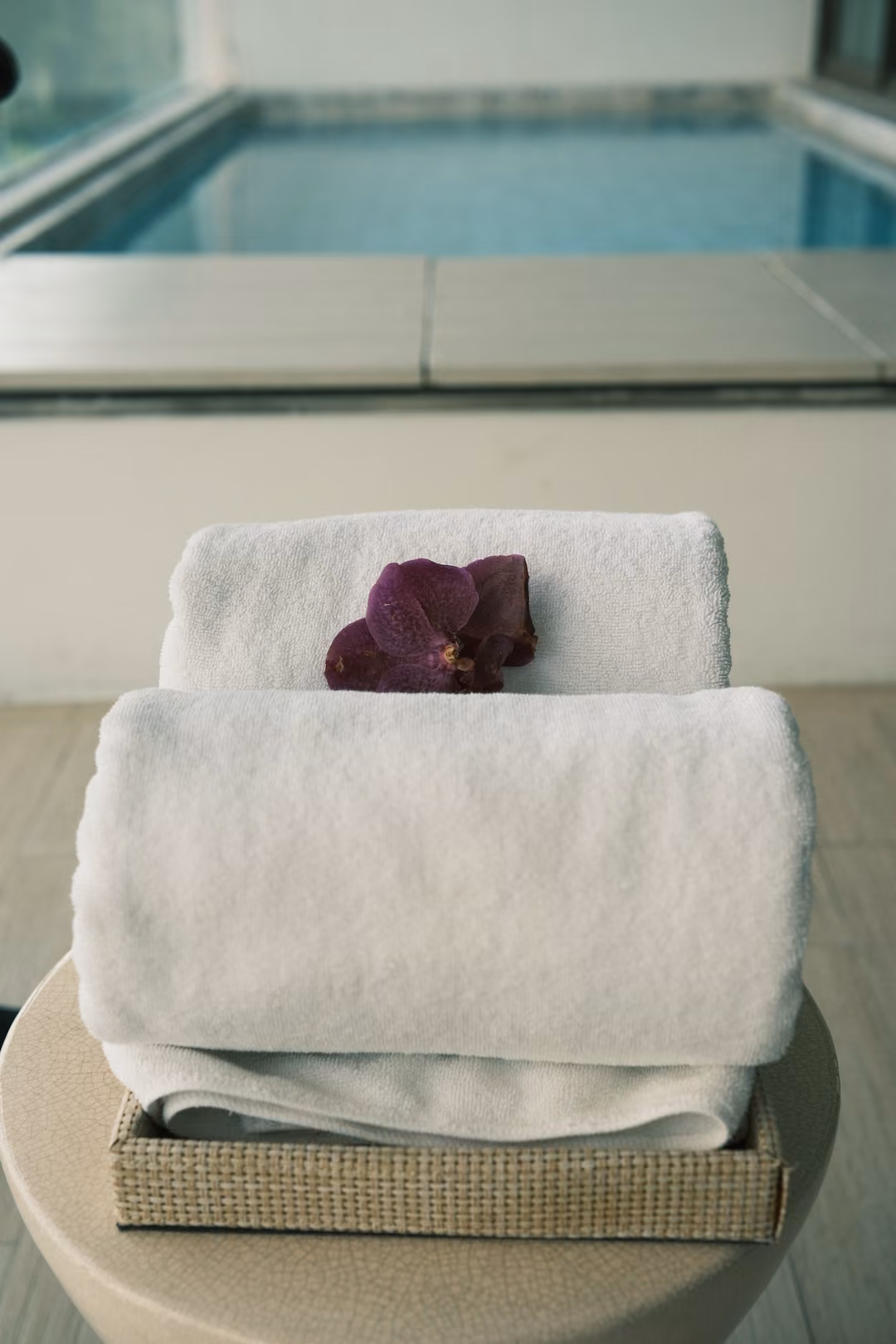 Rolled white towels topped with a purple orchid on a small table, a pool blurred behind