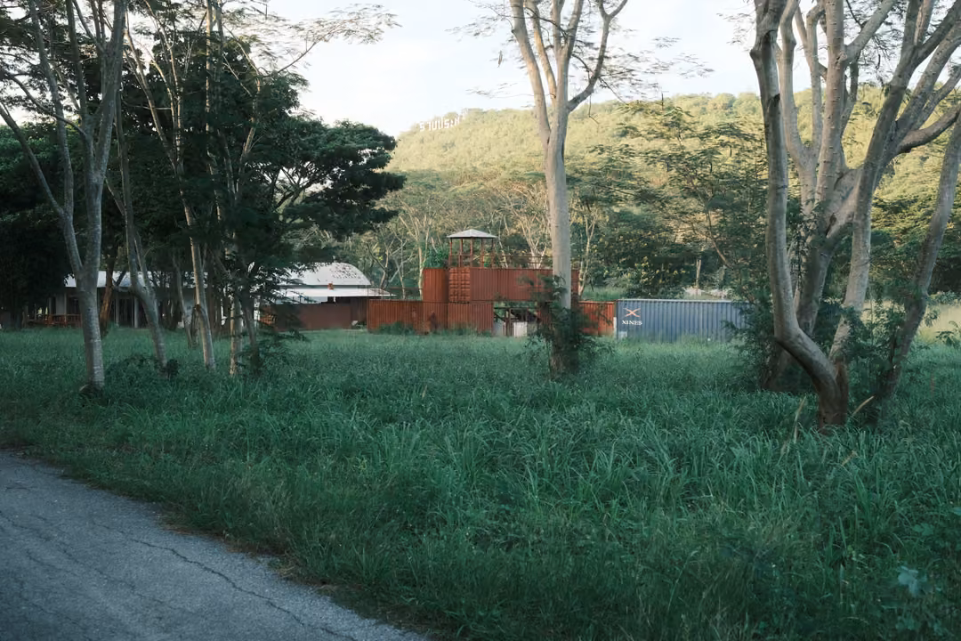Grassy field with scattered trees and rust-red shipping containers stacked into structures, green hills behind