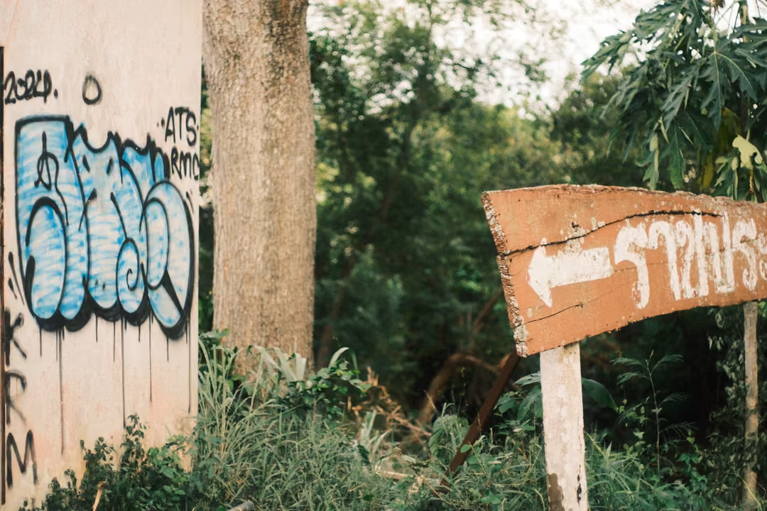 A concrete wall with blue graffiti beside a weathered wooden arrow sign with Thai lettering, trees behind.