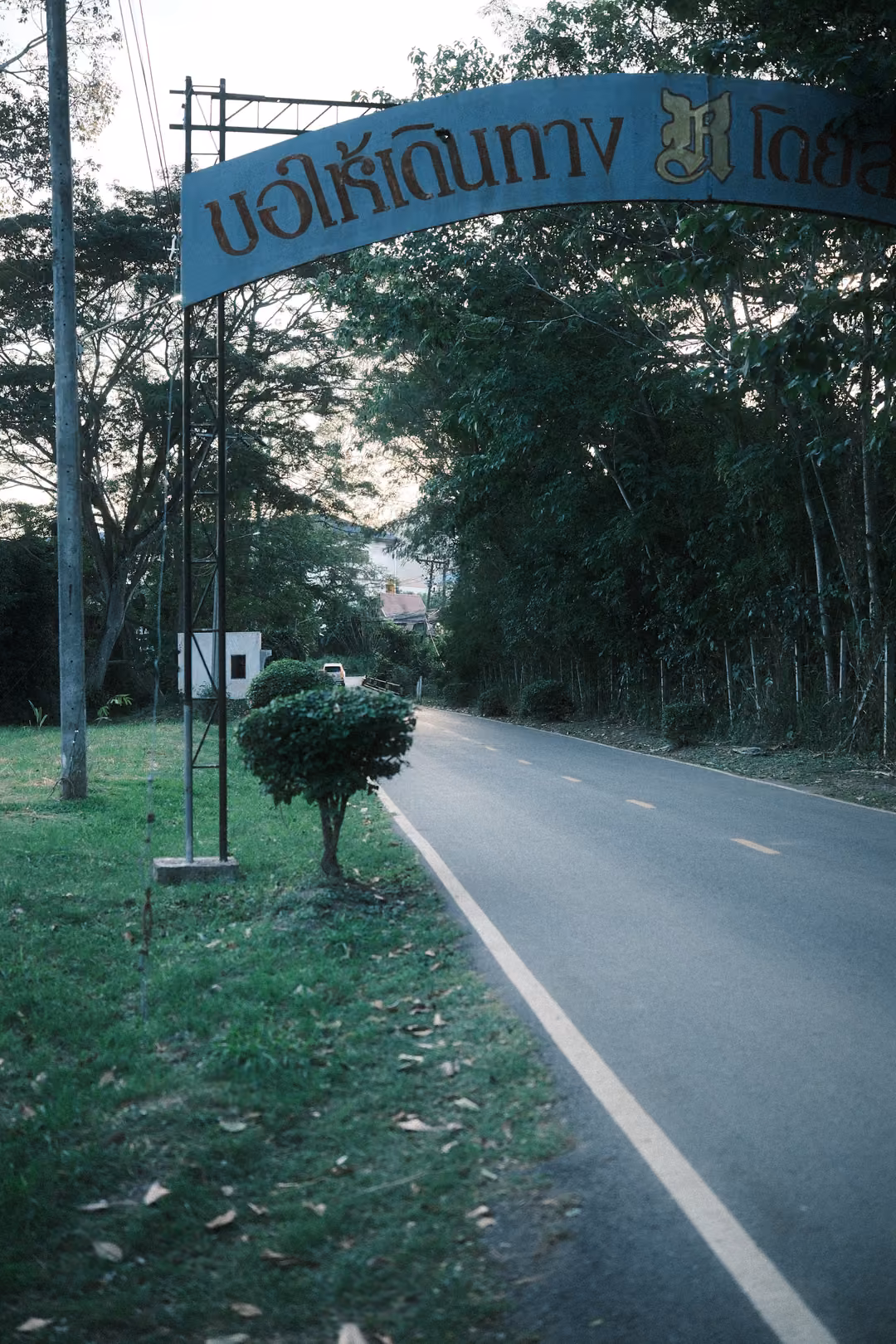 Blue arched roadside sign with Thai text over a grassy verge, a road with white lines leading into trees