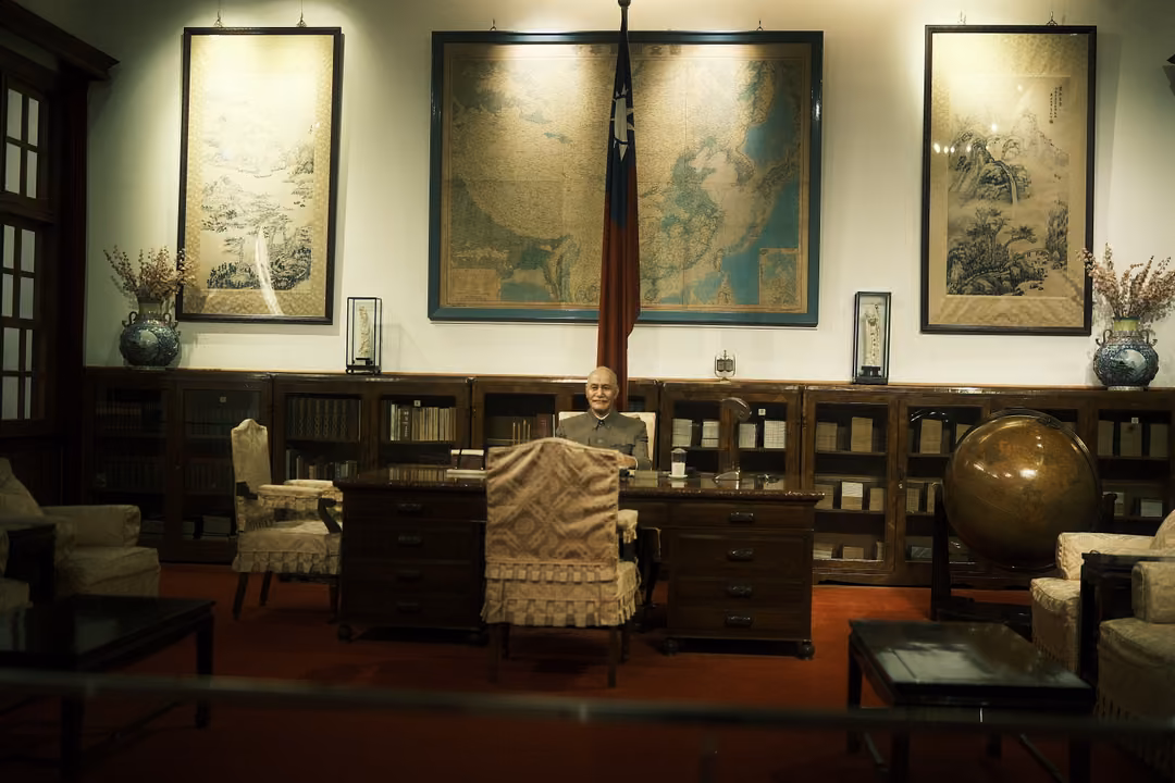 Recreated office with a seated figure at a wooden desk, maps and scroll paintings on the wall, a flag in the corner