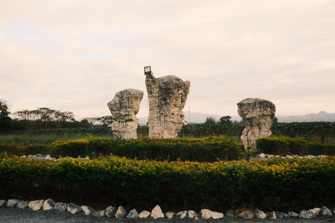 Three tall rough stone pillars in a garden ringed by hedges and white stones, cloudy sky and trees behind