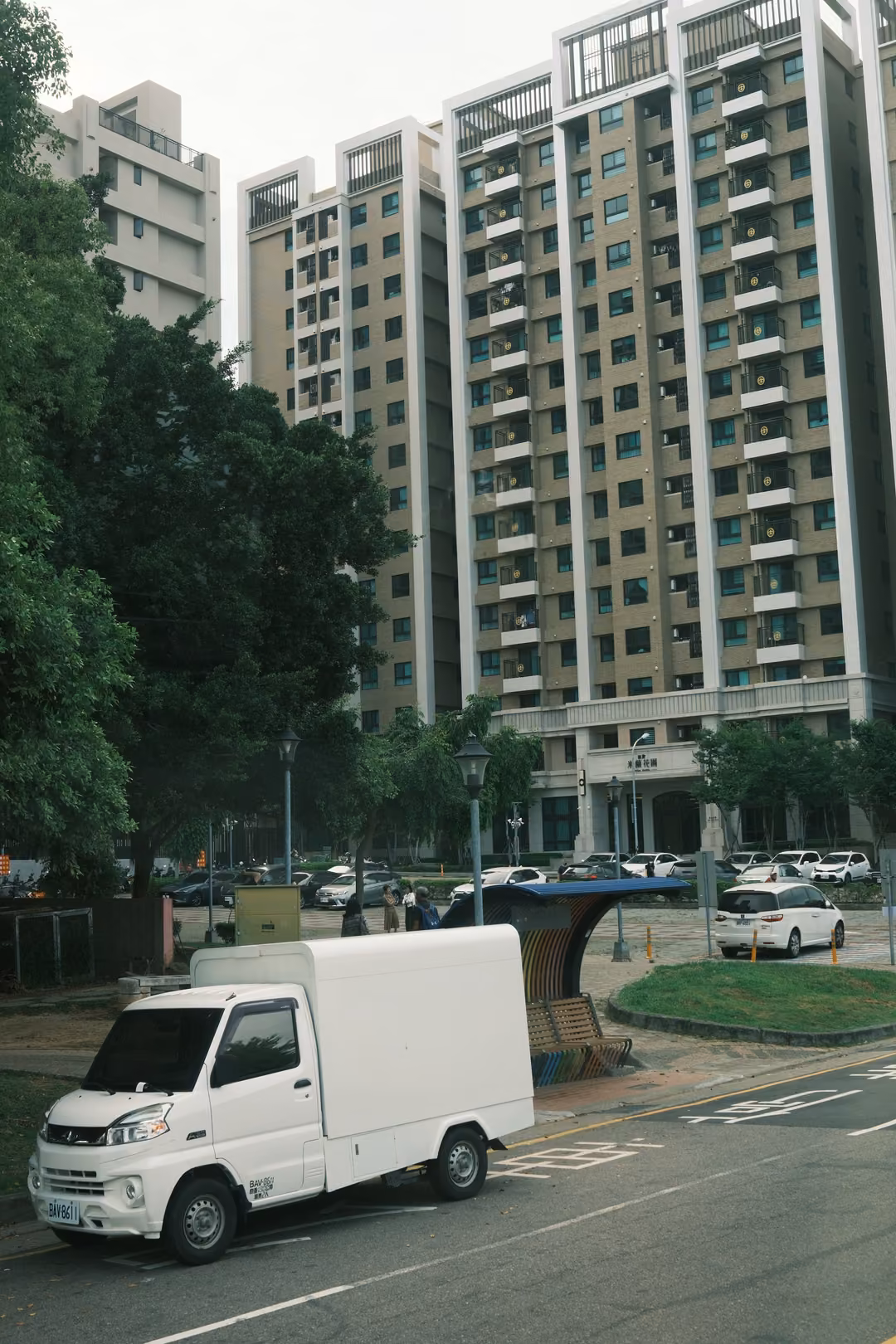 White mini delivery truck parked on a street below tall residential towers and green trees