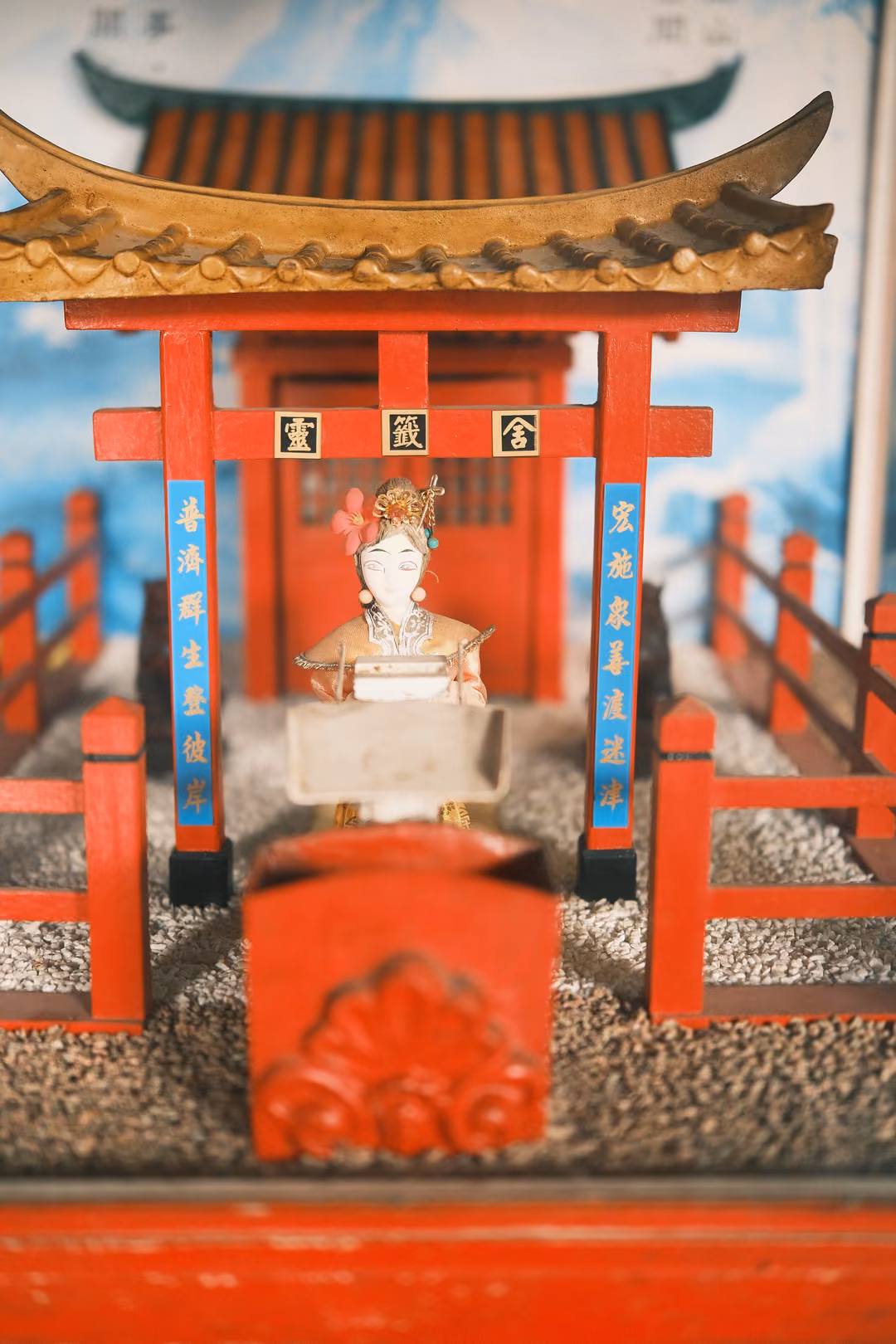 Miniature red shrine with a curved roof and a small crowned figure, set on gravel behind a red railing