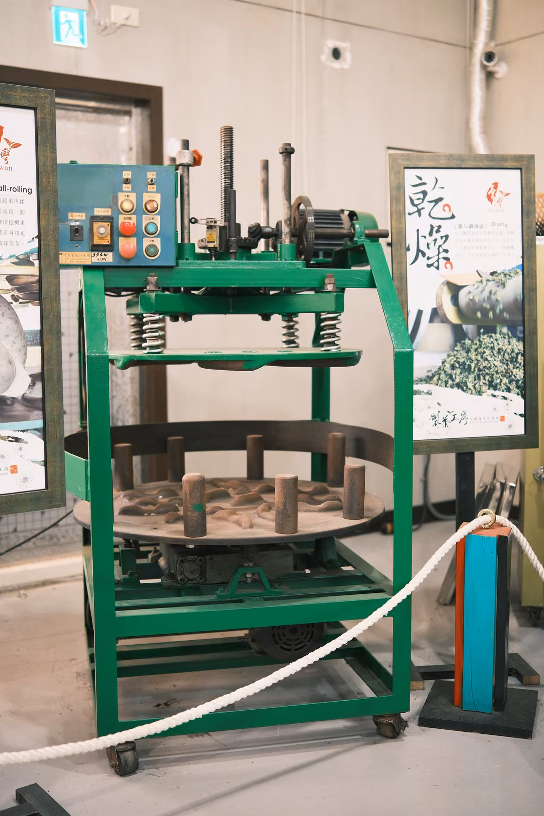 Green metal tea-rolling machine with a round tray and wooden pegs, roped off beside a poster reading 乾燥
