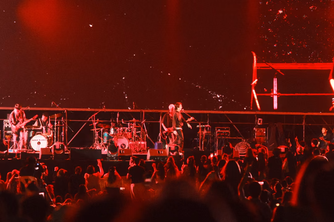 Stage and crowd bathed in deep red light, a band performing, white flecks drifting on the screen behind.