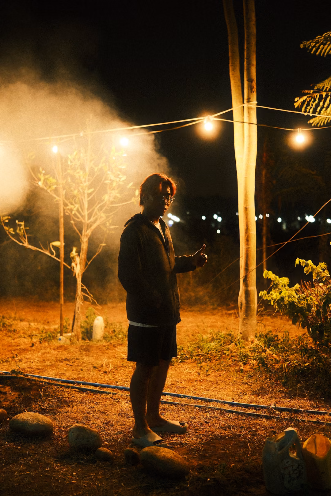 Vertical frame of a person in a dark hoodie and shorts standing barefoot on stones under smoky string lights