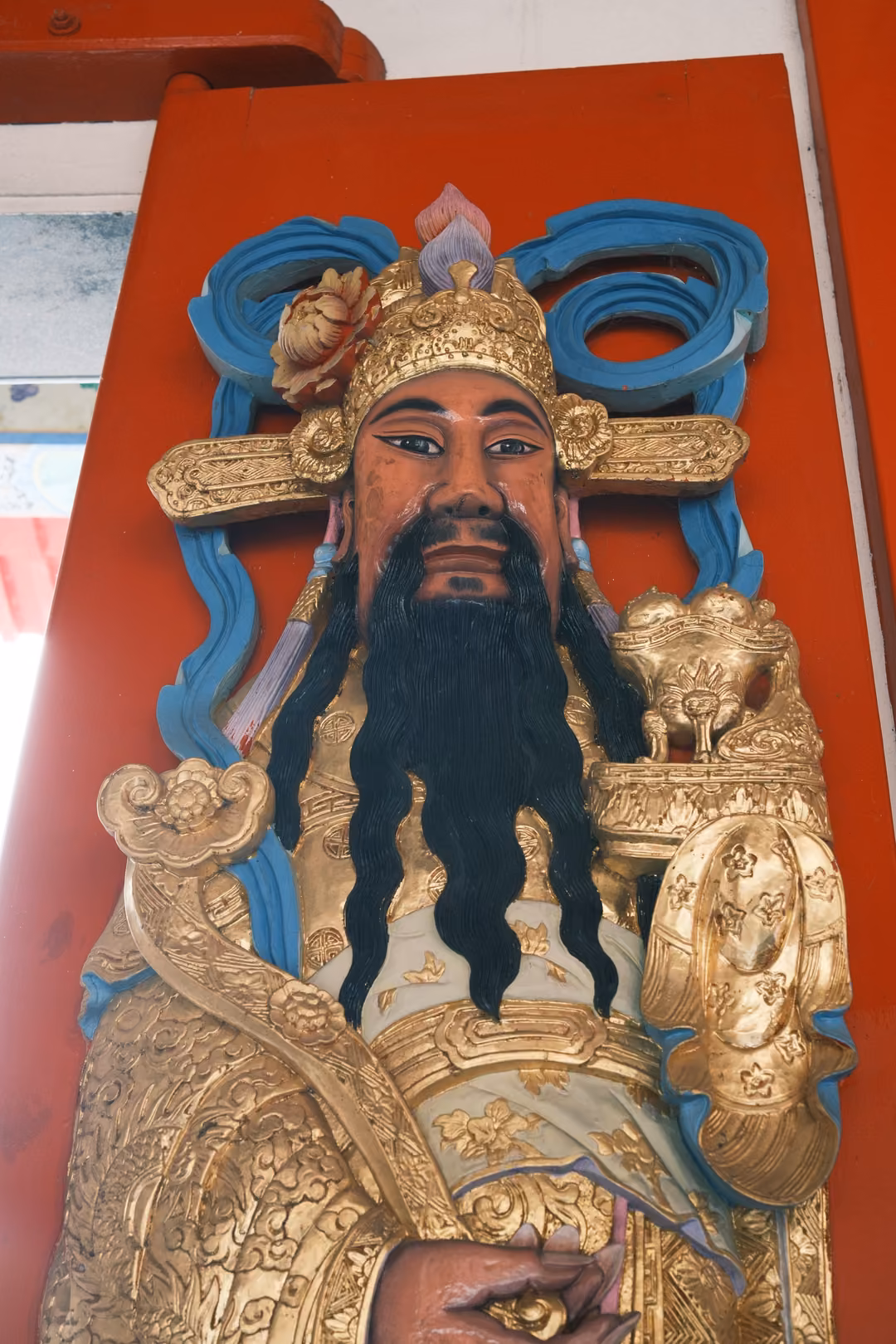 Painted statue of a bearded figure with gold crown and armour, blue ribbons, against a red pillar