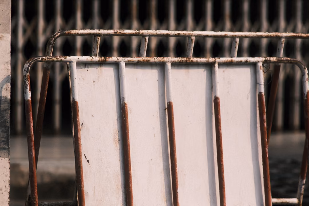 A white metal crowd barrier with rusted bars, in front of a dark folding metal gate.