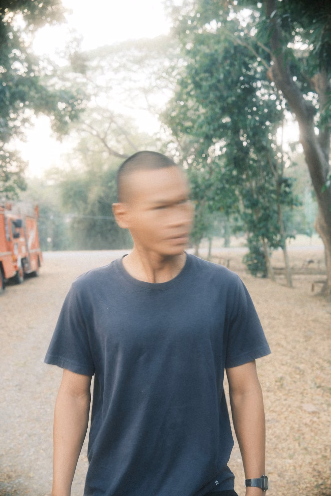Man in a grey t-shirt with motion-blurred face turning to the right, trees and red vehicle behind