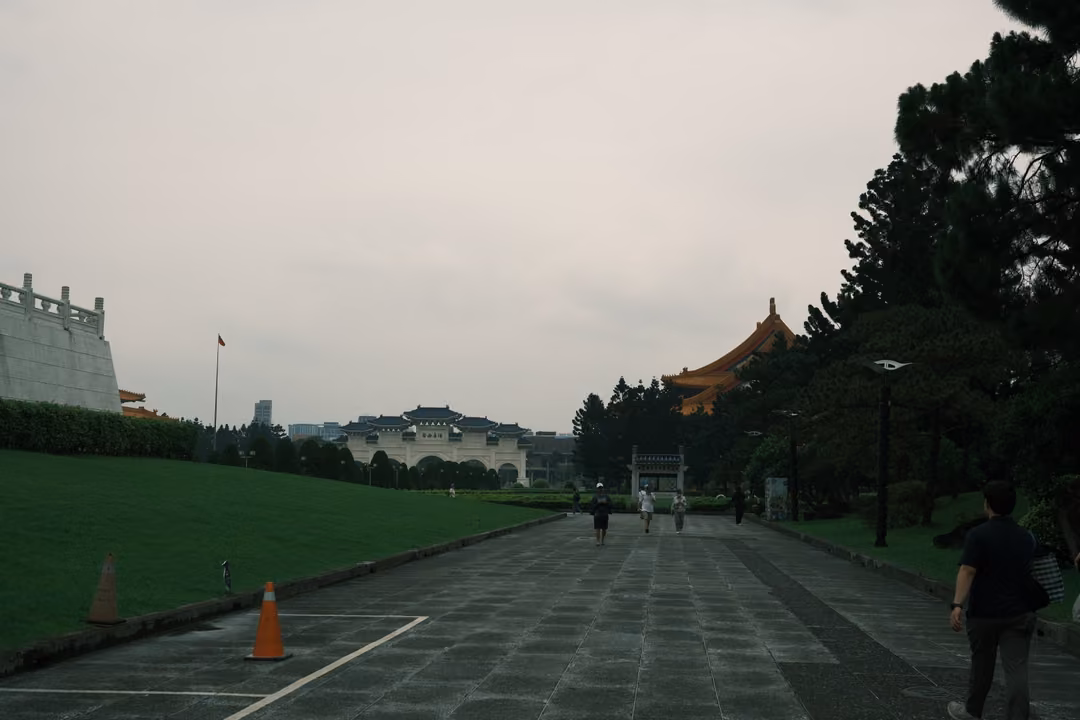 Same long avenue toward a distant gate, a person walking at right, cones on the paving