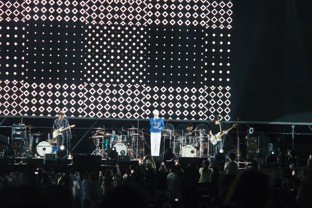 Band on stage below a screen of white diamond patterns in rectangles, the crowd visible in the foreground.