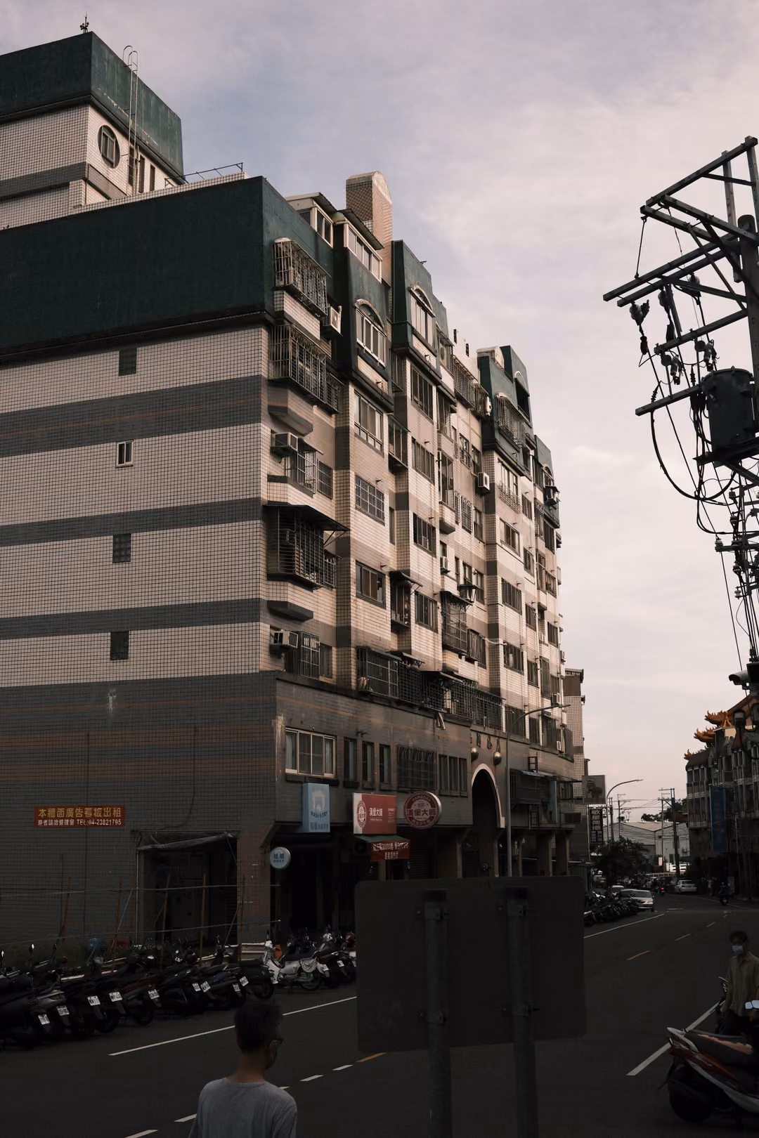 Tall apartment building beside a utility pole with transformers, scooters parked along the street below