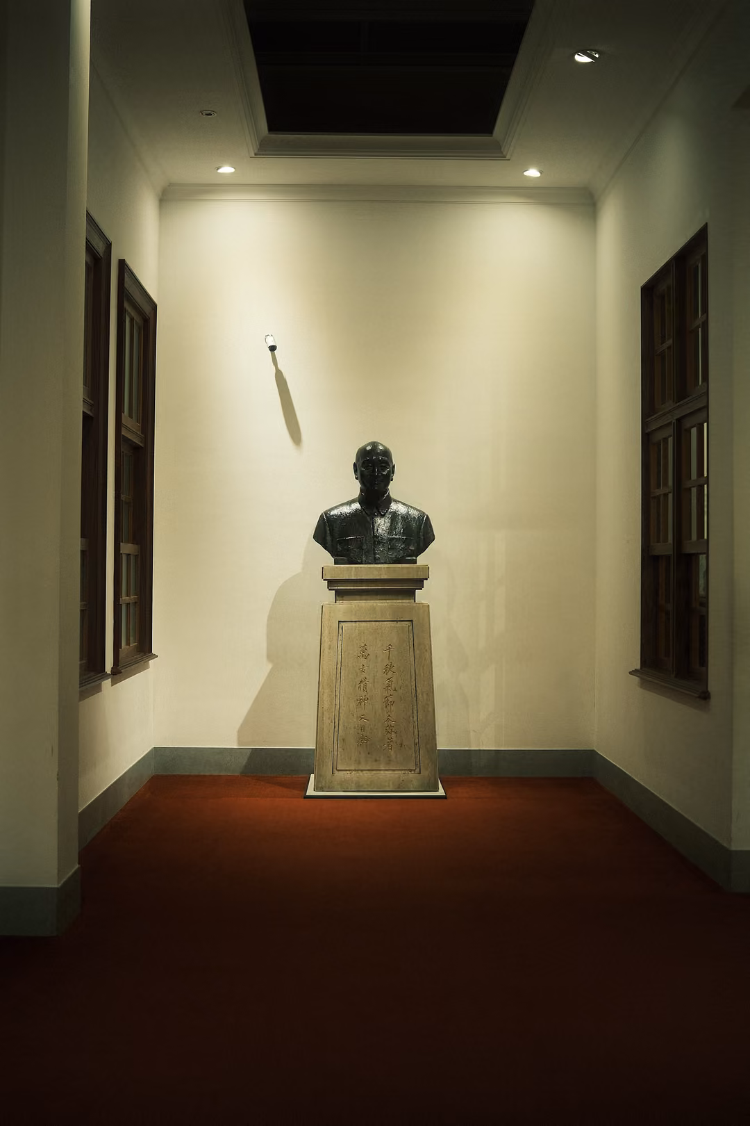 Bronze bust on a stone pedestal at the end of a narrow cream-walled alcove with a red carpet