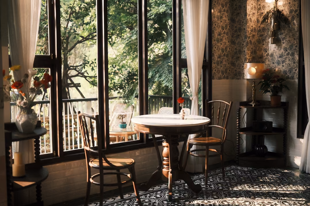 Tall windows onto green trees beside a round table with two wooden chairs on a patterned tile floor