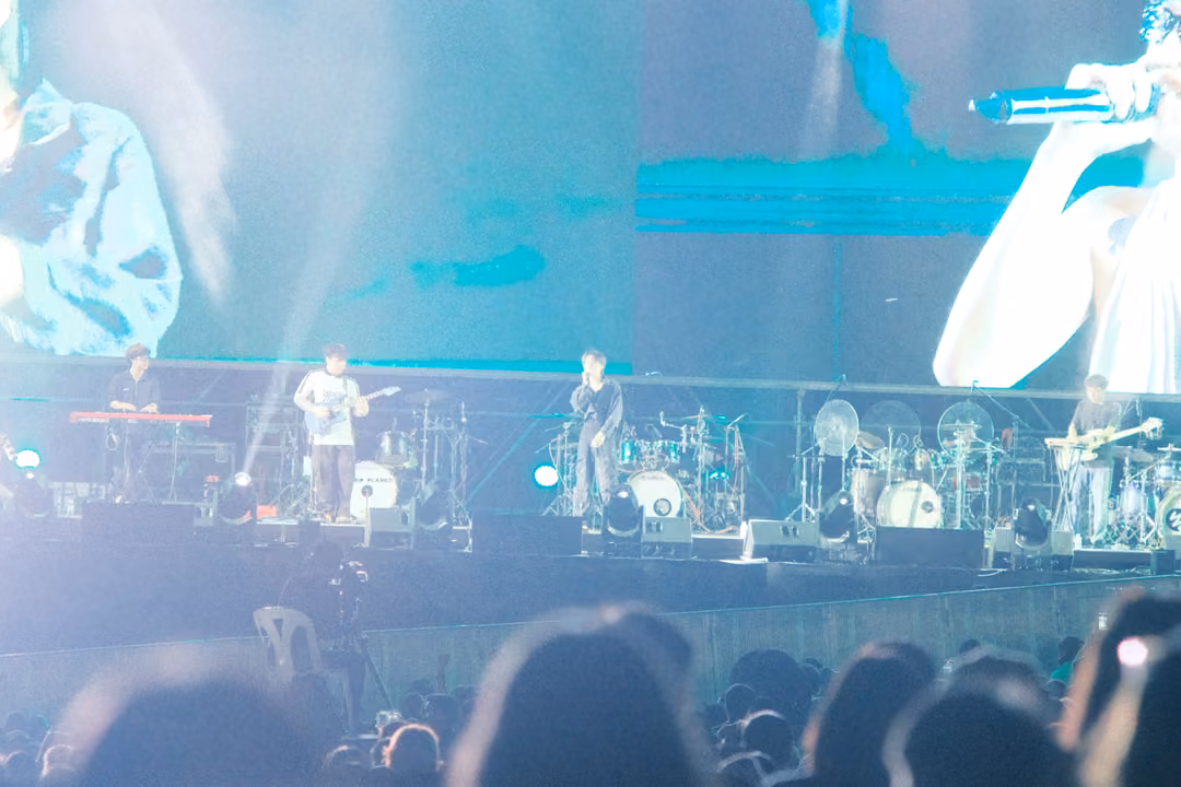 Band on a stage washed in pale cyan light, screen close-ups above, blurred heads of the crowd in the foreground.