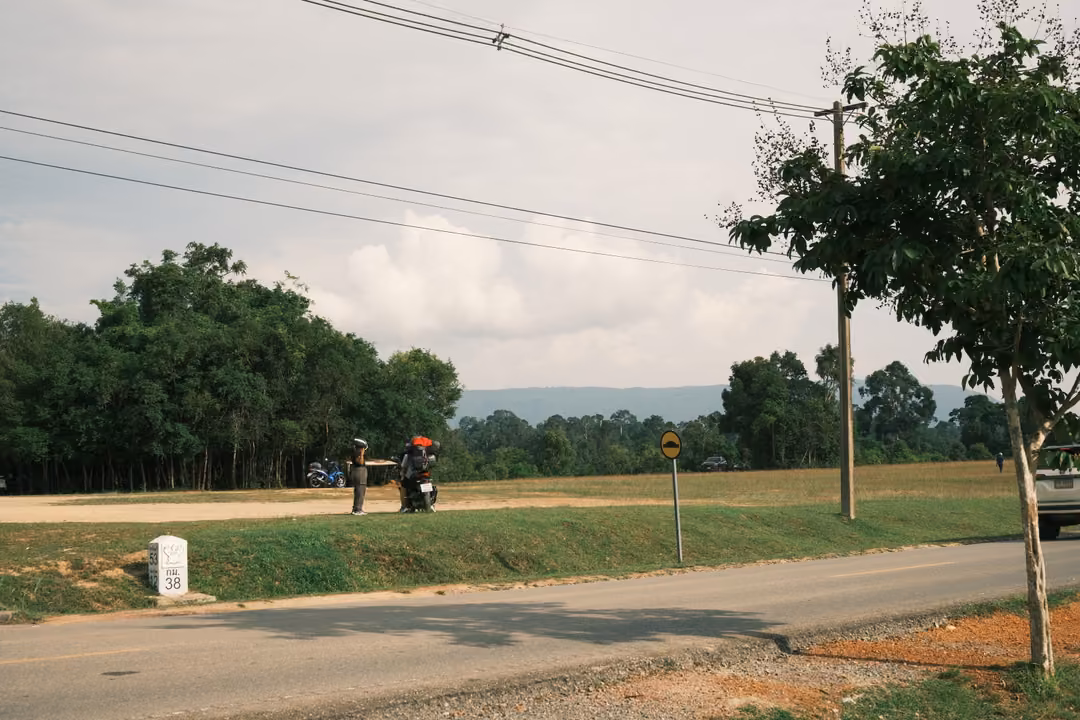 A roadside with a motorbike rider, a white milestone, a small tree and power lines under a cloudy sky.