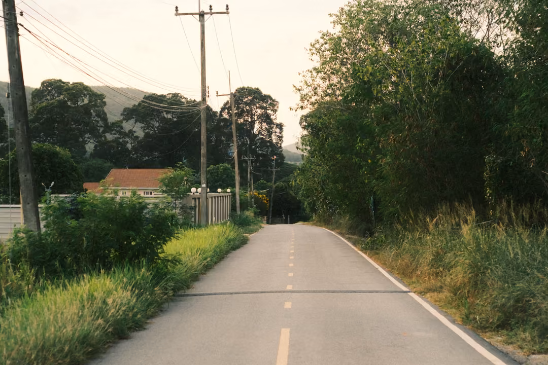 A quiet road with a dashed centre line, power poles and wires, red-roofed houses and hills under a pale sky.