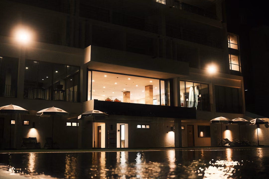 Large hotel building at night with lit glass lobby, white umbrellas and lights reflected in a pool