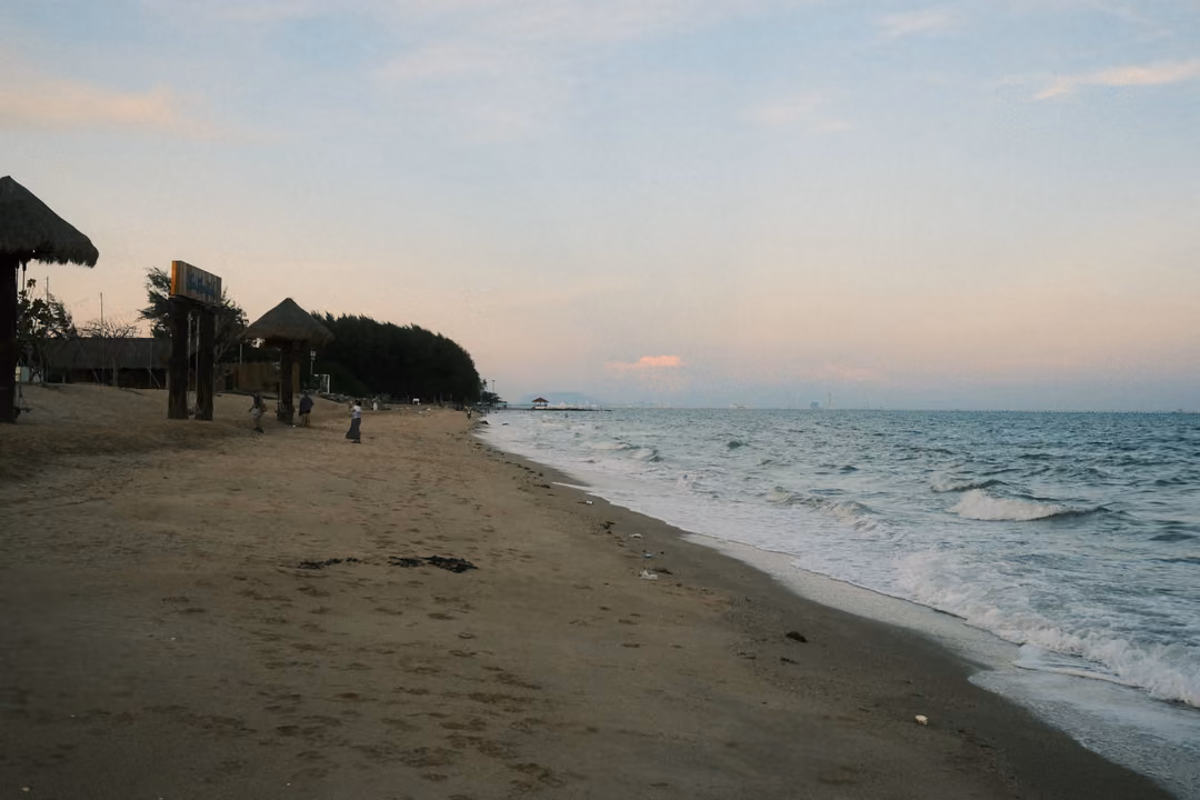 Long sandy beach curving into the distance at dusk, thatched umbrellas and trees, waves washing in