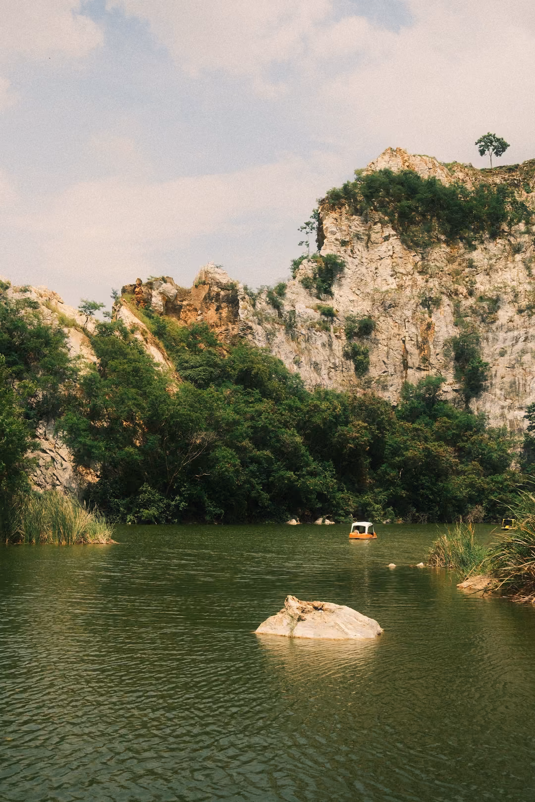 Vertical view of a green lake with a pale rock and pedal boat, cliffs and trees under a cloudy blue sky