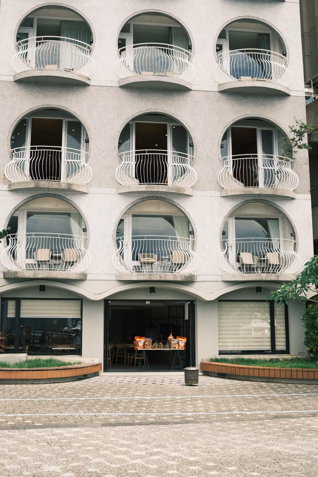 White building with arched windows and curved white balconies above a tiled plaza and planters