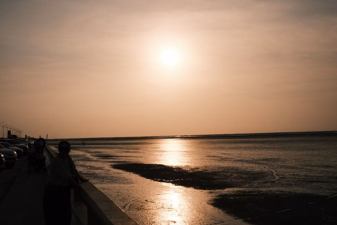 A low sun in a peach sky over mudflats, figures silhouetted along a railing in the foreground