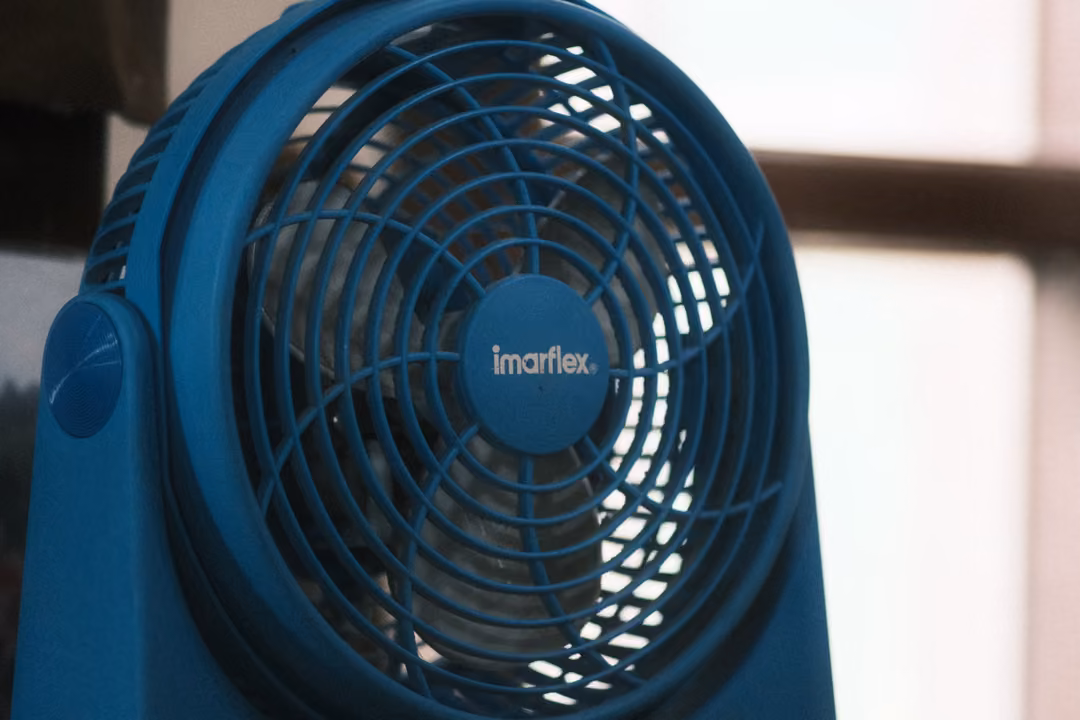 A blue Imarflex desk fan with a round grille in front of a pale wall and window frame.