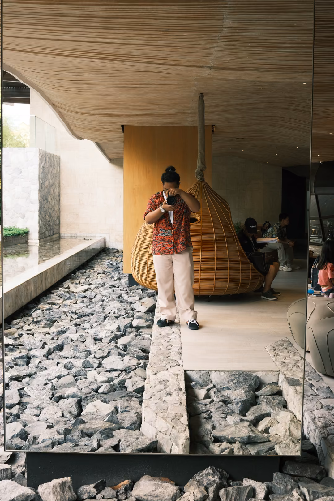 A figure in a red shirt reflected in a mirror in an open lobby with stone floors and a hanging rattan seat