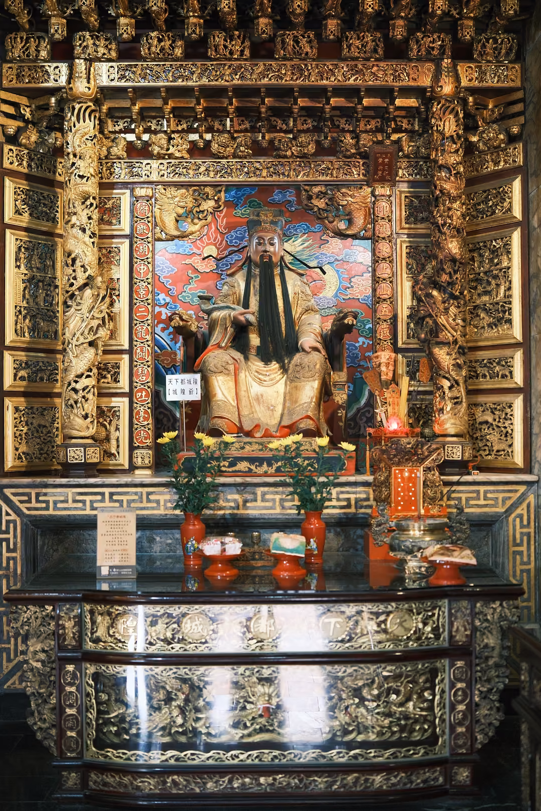 Ornate gilded temple altar with a seated bearded figure in red and gold robes, flowers and offerings below