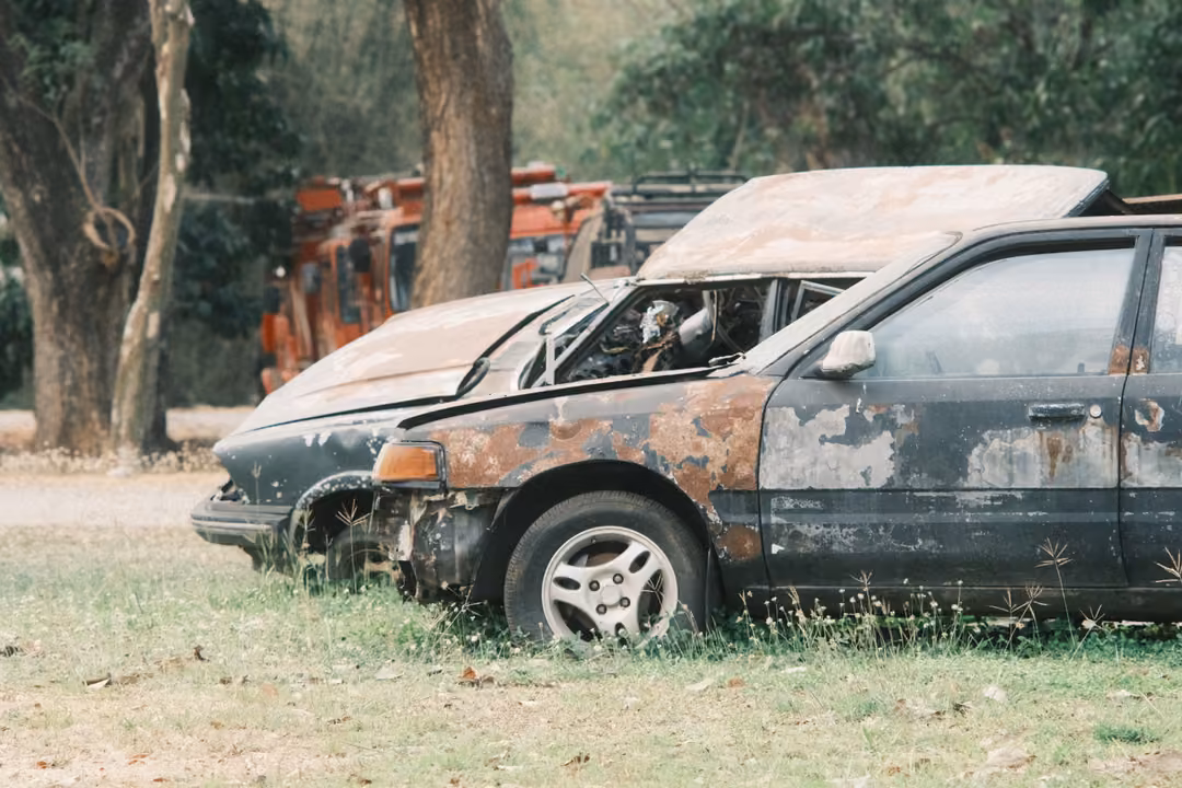 Burned, rusted car wreck in the grass with a red vehicle and trees behind