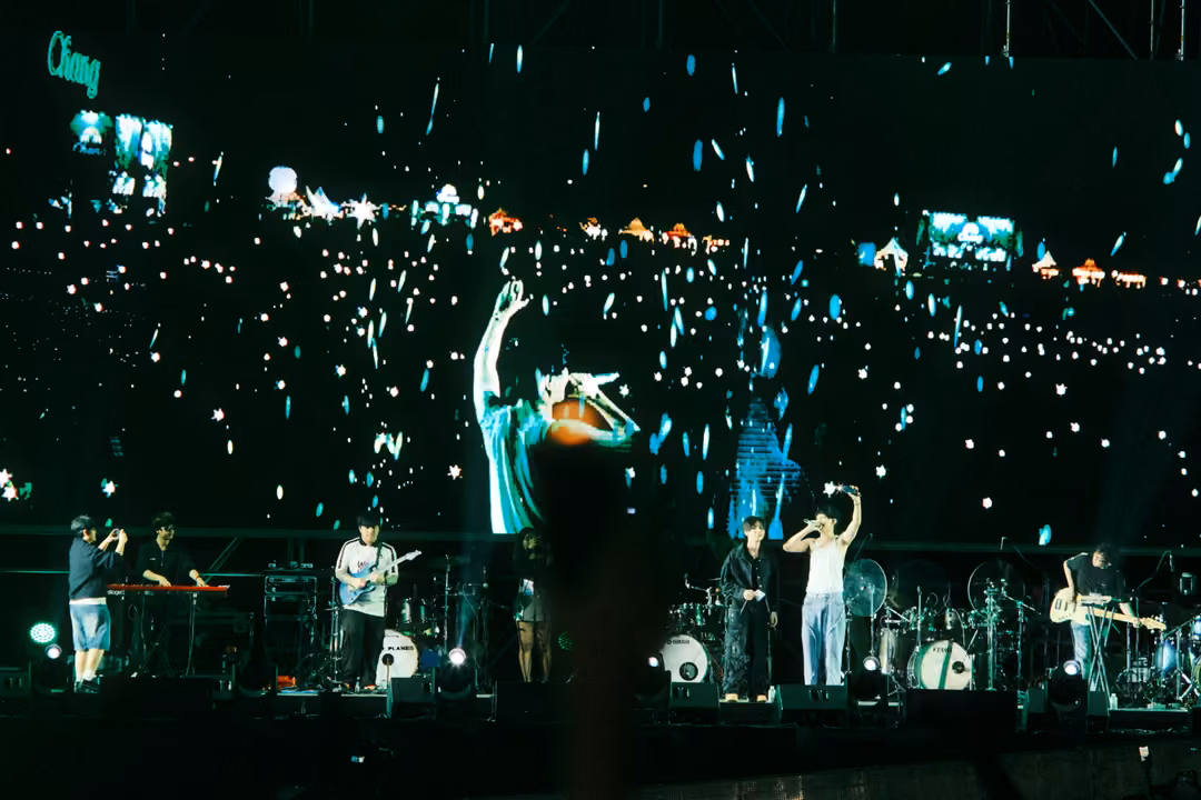 Screen showing a singer raising one arm toward a field of phone lights, band on stage below, blurred hands in front.