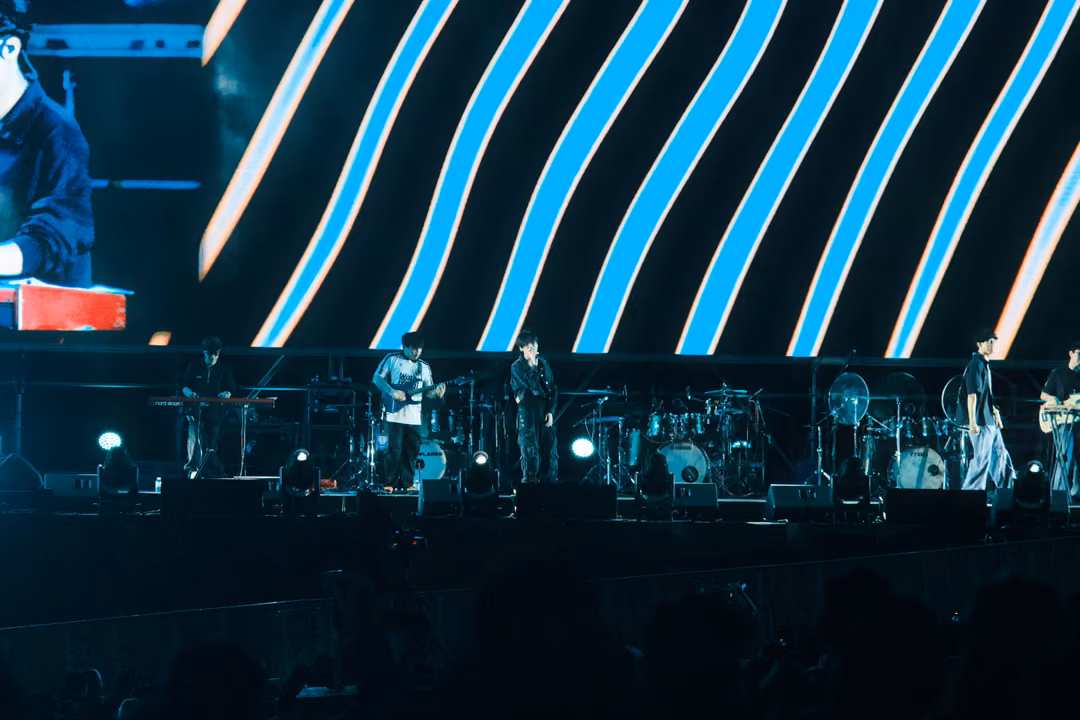 Wide stage view with blue and white diagonal stripes on the screen and the band spread across a lit platform.
