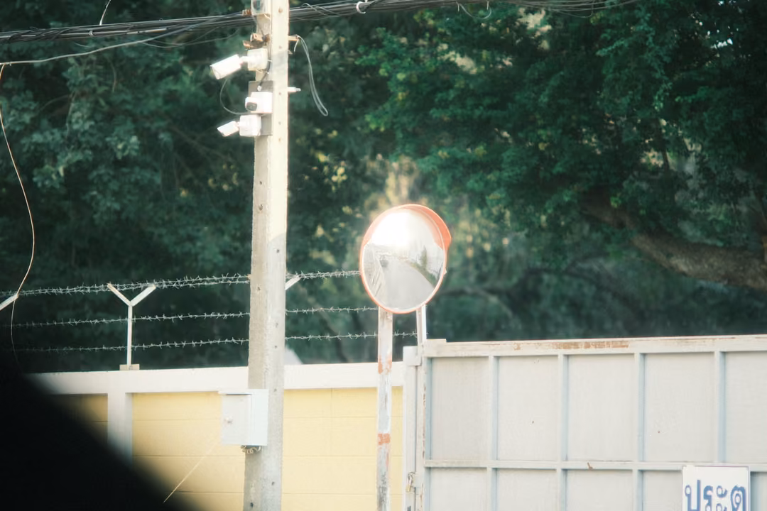 Convex road mirror on a pole beside a wall topped with barbed wire, security cameras and trees
