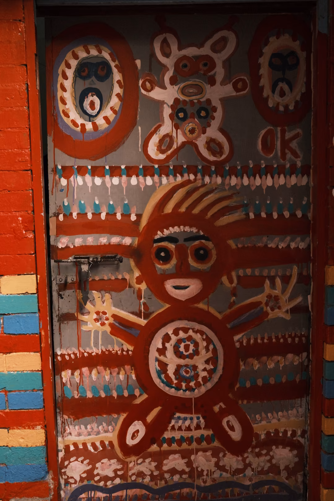 Red door painted with a white wide-eyed figure, masks, a bear shape and the letters OK beside striped bricks